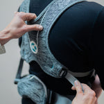 A person wearing the Carifit+ Baby Carrier in Cool Grey. The image shows the back on the carrier on the persons back, with another adult adjusting the adaptive sizing straps on the side