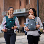 Two adults carrying a child in the Carifit+ Fabric Baby Carrier in Cool Grey and Cool Green with a town/city scape scene behind them