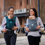 Two adults carrying a child in the Carifit+ Fabric Baby Carrier in Cool Grey and Cool Green with a town/city scape scene behind them