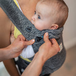 A child sat in the Carifit+ Baby Carrier in Cool Grey with an adult showing the removeable head neck support which attaches onto the front of the carrier via two clips on the carrier, and elasticated loops on the head and neck support