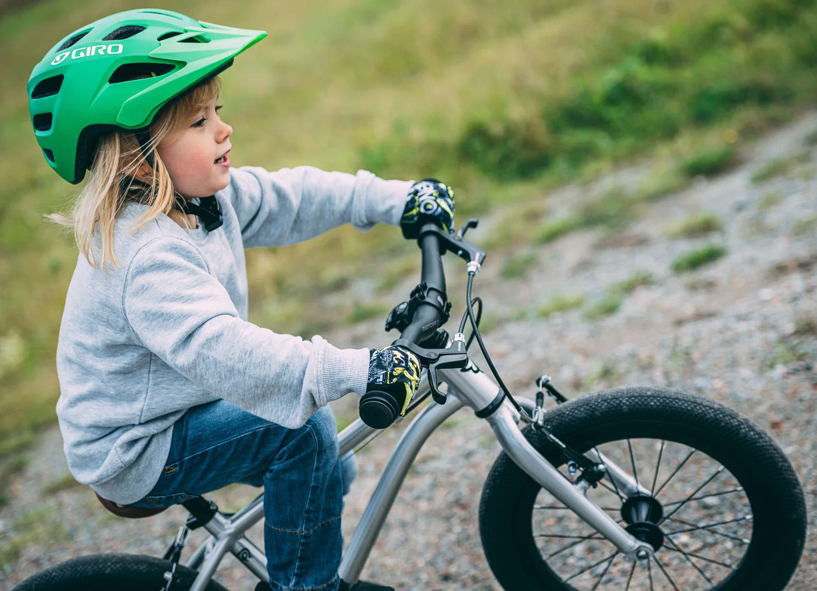 Child wearing a green helmet and gray jacket riding Early Rider's Belter 16 inch wheel children's bike on a gravel path.