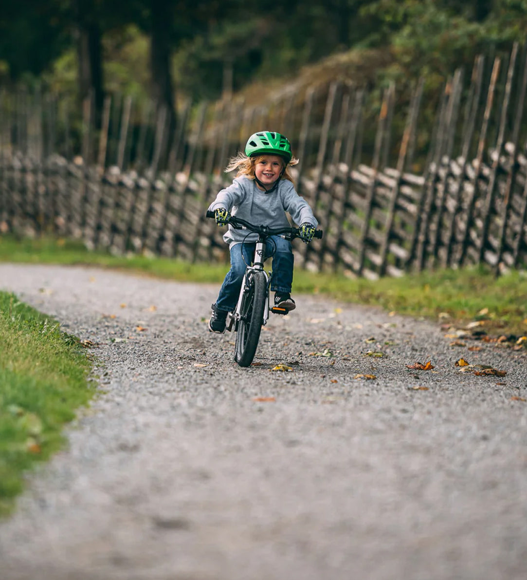 Child smiling while riding Early Rider's Belter 16 inch wheel children's bike on a gravel path with a wooden fence and trees in the background.