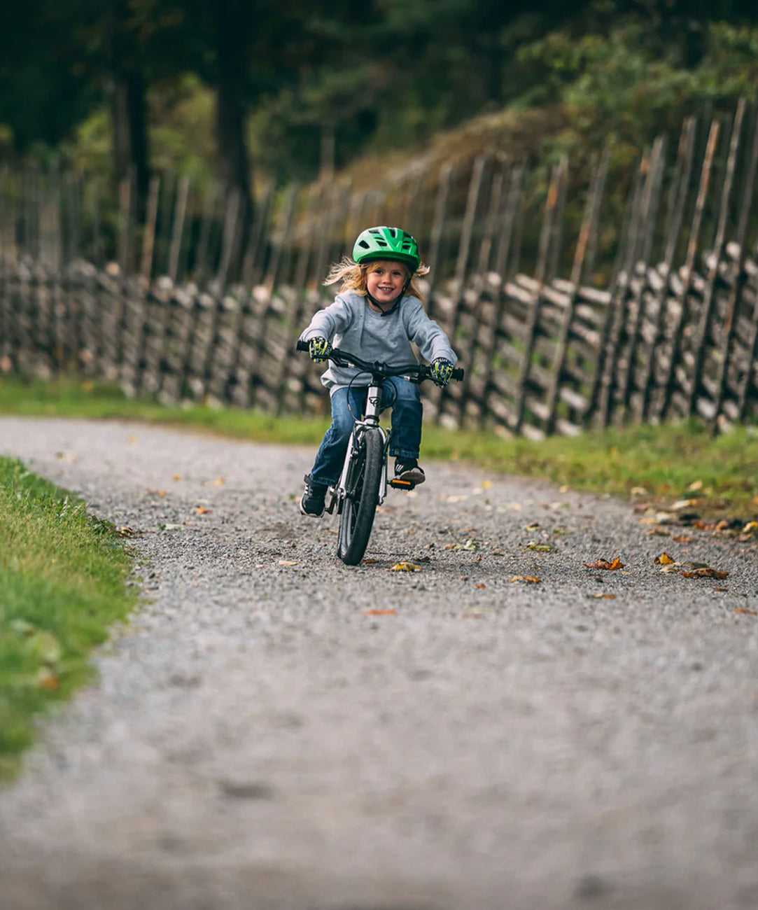 Child smiling while riding Early Rider's Belter 16 inch wheel children's bike on a gravel path with a wooden fence and trees in the background.