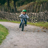 Child smiling while riding Early Rider's Belter 16 inch wheel children's bike on a gravel path with a wooden fence and trees in the background.