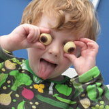 Cocoa Loco chocolate eyeballs being held by a child. The child is sticking his tongue out and is holding the chocolate eyeballs against his eyes. He is wearing a black top with a vegetable print on it.