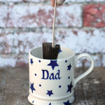 Cocoa Loco hot chocolate spoon in a hand hovering over a white ceramic mug. The mug has a blue star pattern on it and the words dad in the middle.