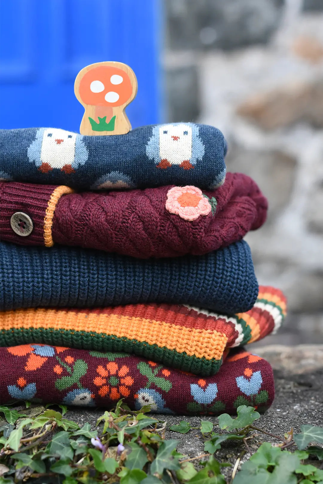 A stack of colorful organic cotton and merino wool kids' knitted sweaters with whimsical patterns, animals, flowers, and stripes, topped with a small  wooden Lanka Kade x babipur mushroom-shaped toy, set outdoors on a stone surface with green leaves.