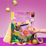 A hand holds a small packaged item in front of a colorful Divine Chocolate brand advent calendar with Christmas-themed illustrations, set on a purple surface against a pink-purple background with hanging gold ornaments.