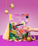 A hand holds a small packaged item in front of a colorful Divine Chocolate brand advent calendar with Christmas-themed illustrations, set on a purple surface against a pink-purple background with hanging gold ornaments.