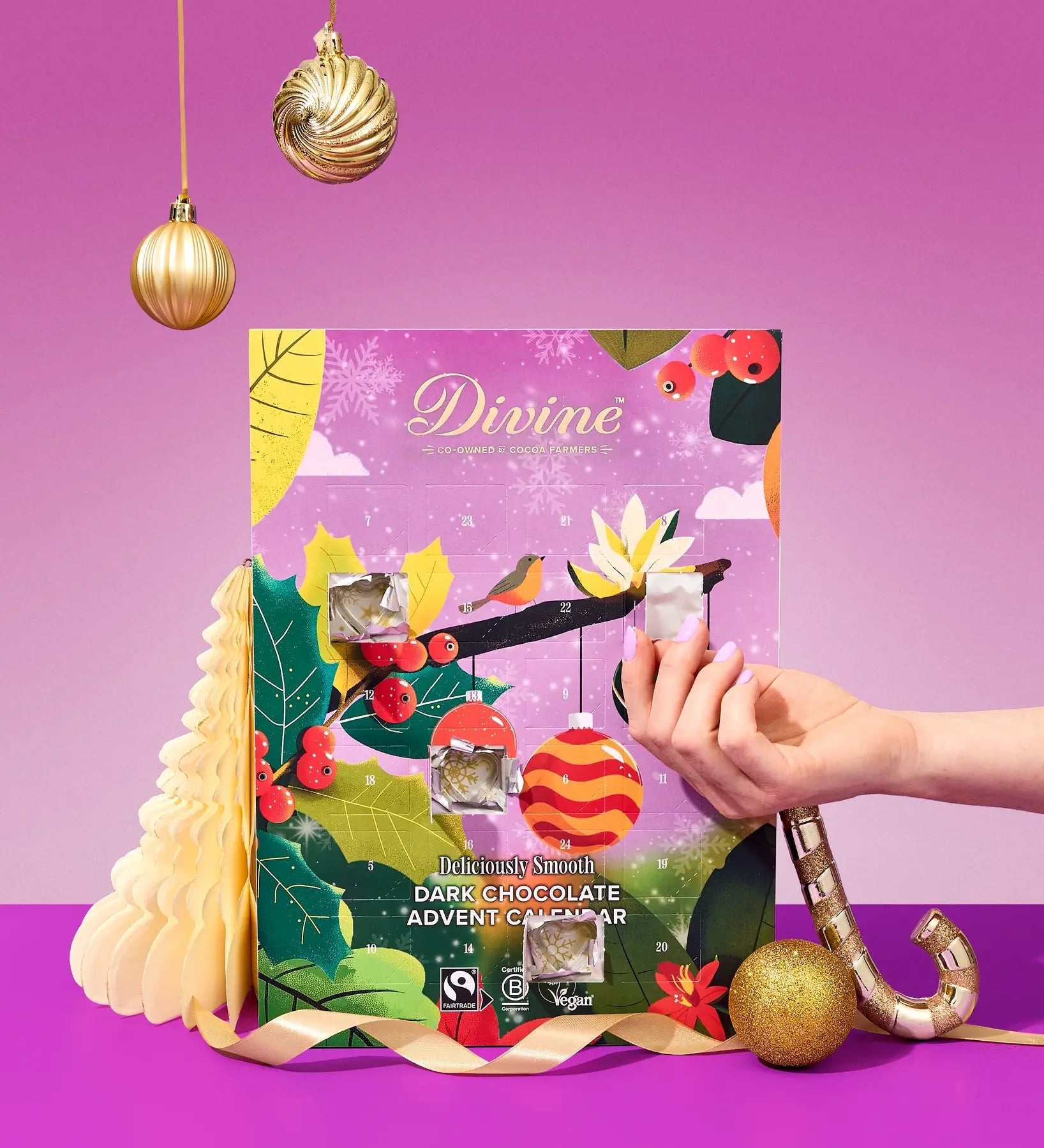 A hand holds a small packaged item in front of a colorful Divine Chocolate brand advent calendar with Christmas-themed illustrations, set on a purple surface against a pink-purple background with hanging gold ornaments.