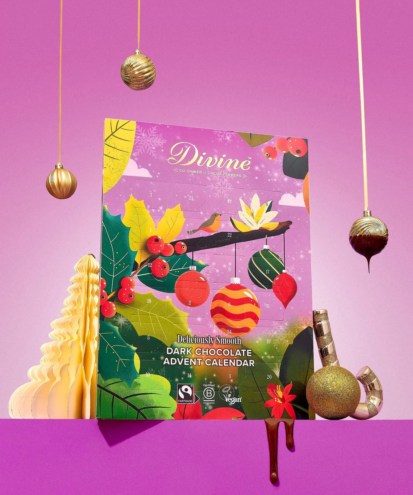 A decorative product shot of a Divine Chocolate themed Advent Calendar, surrounded by festive props including hanging gold ornaments, a gold Christmas tree, chocolate drizzle, and wrapped chocolates on a solid pink background.