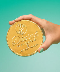 A hand with mint green nails holds a giant round gold-colored Divine Chocolate coin with milk chocolate against a solid teal background.