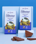 2 Divine fairtrade 90g smooth milk chocolate bars standing against a blue platform with loose chocolate pieces in front