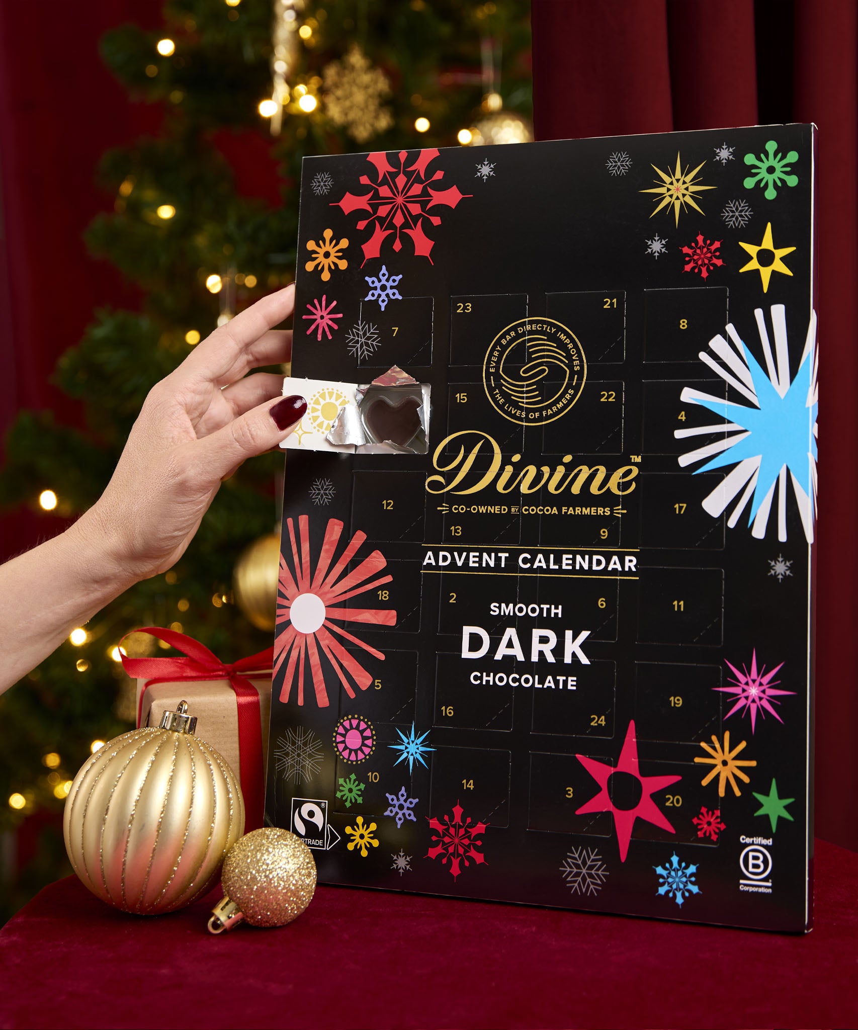 Divine fairtrade dark chocolate advent calendar available at Babipur. Showing the front with an adult hand opening one of the windows. Calendar is standing in a festive inspired display.