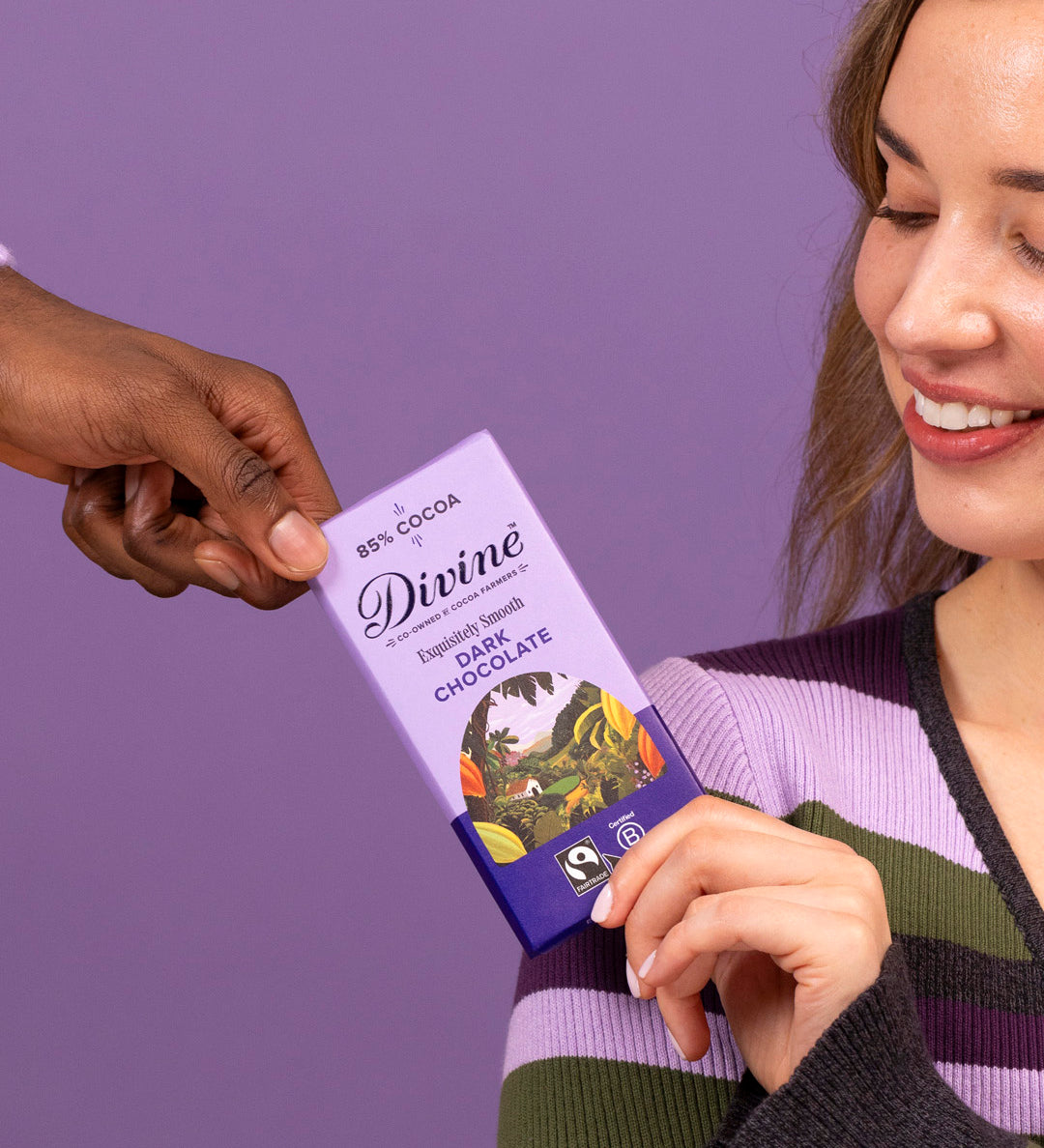 2 adults holding a Divine fairtrade smooth dark chocolate bar showing the purple wrapping with a cocoa farm illustration 