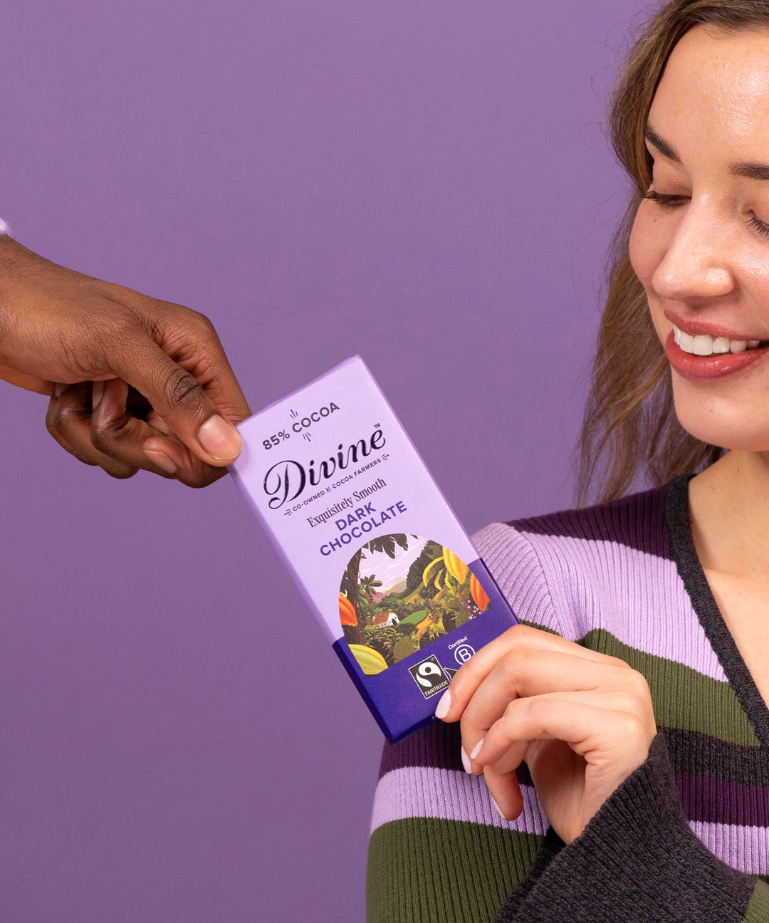 2 adults holding a Divine fairtrade smooth dark chocolate bar showing the purple wrapping with a cocoa farm illustration 