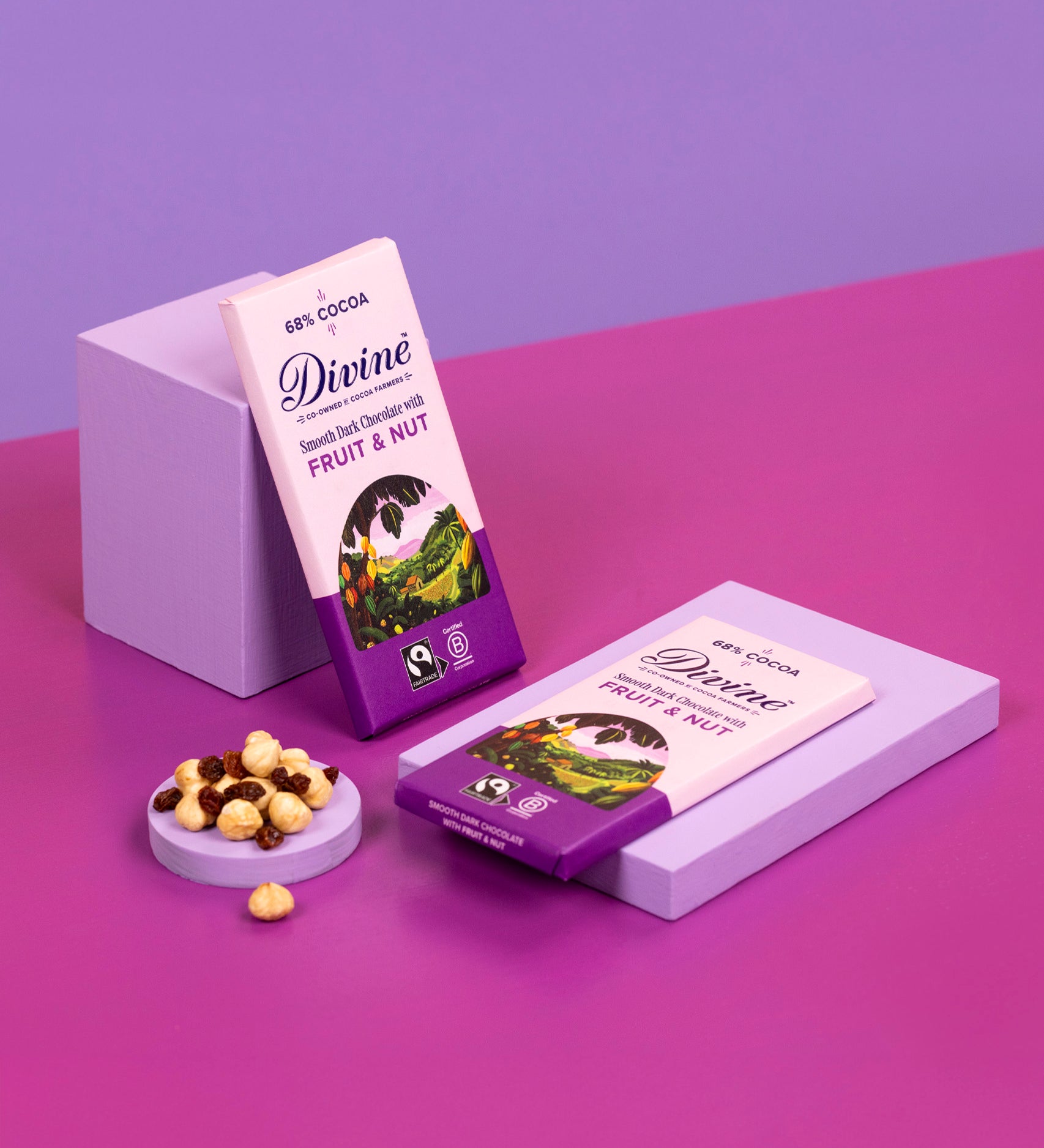 2 bard of Divine fairtrade smooth dark chocolate bar with fruit and nut resting on purple blocks with loose nuts and raisins