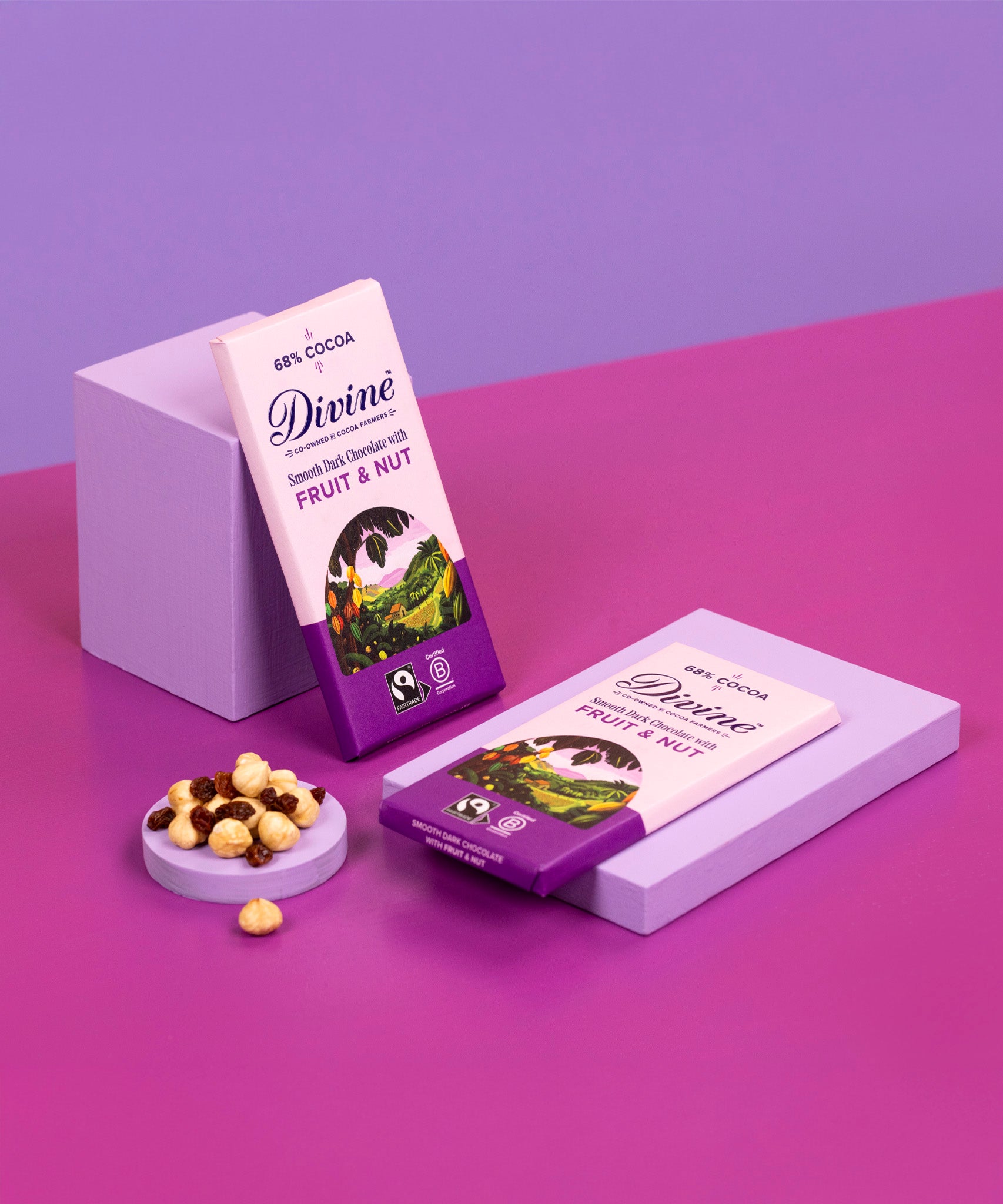 2 bard of Divine fairtrade smooth dark chocolate bar with fruit and nut resting on purple blocks with loose nuts and raisins