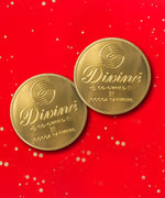 A pair of Divine fairtrade giant milk chocolate coins available at Babipur laid flat with a red festive inspired background.