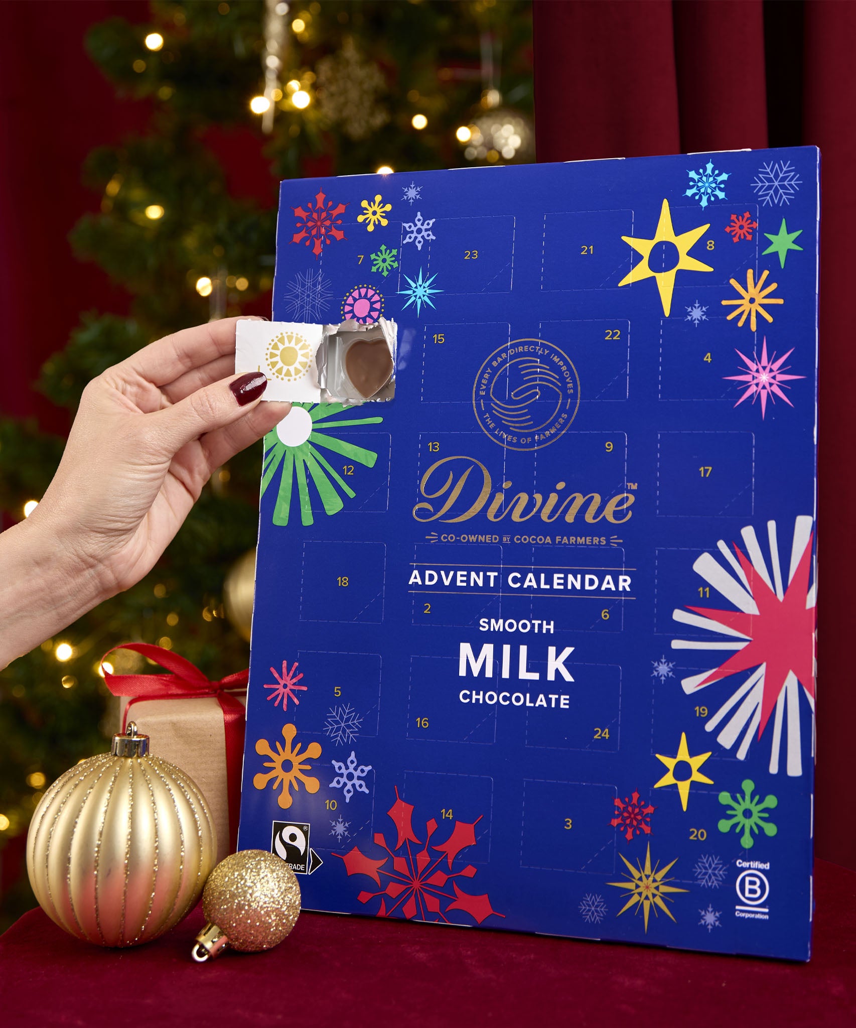 Divine fairtrade milk chocolate advent calendar available at Babipur. Showing the front with an adult hand opening one of the windows. Calendar is standing in a festive display.