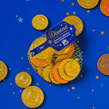 A flatlay with the Divine fairtrade milk chocolate coins inside the yellow netted bag and other coins scattered with a blue festive inspired background. Chocolate coins available at Babipur.