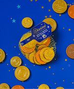 A flatlay with the Divine fairtrade milk chocolate coins inside the yellow netted bag and other coins scattered with a blue festive inspired background. Chocolate coins available at Babipur.