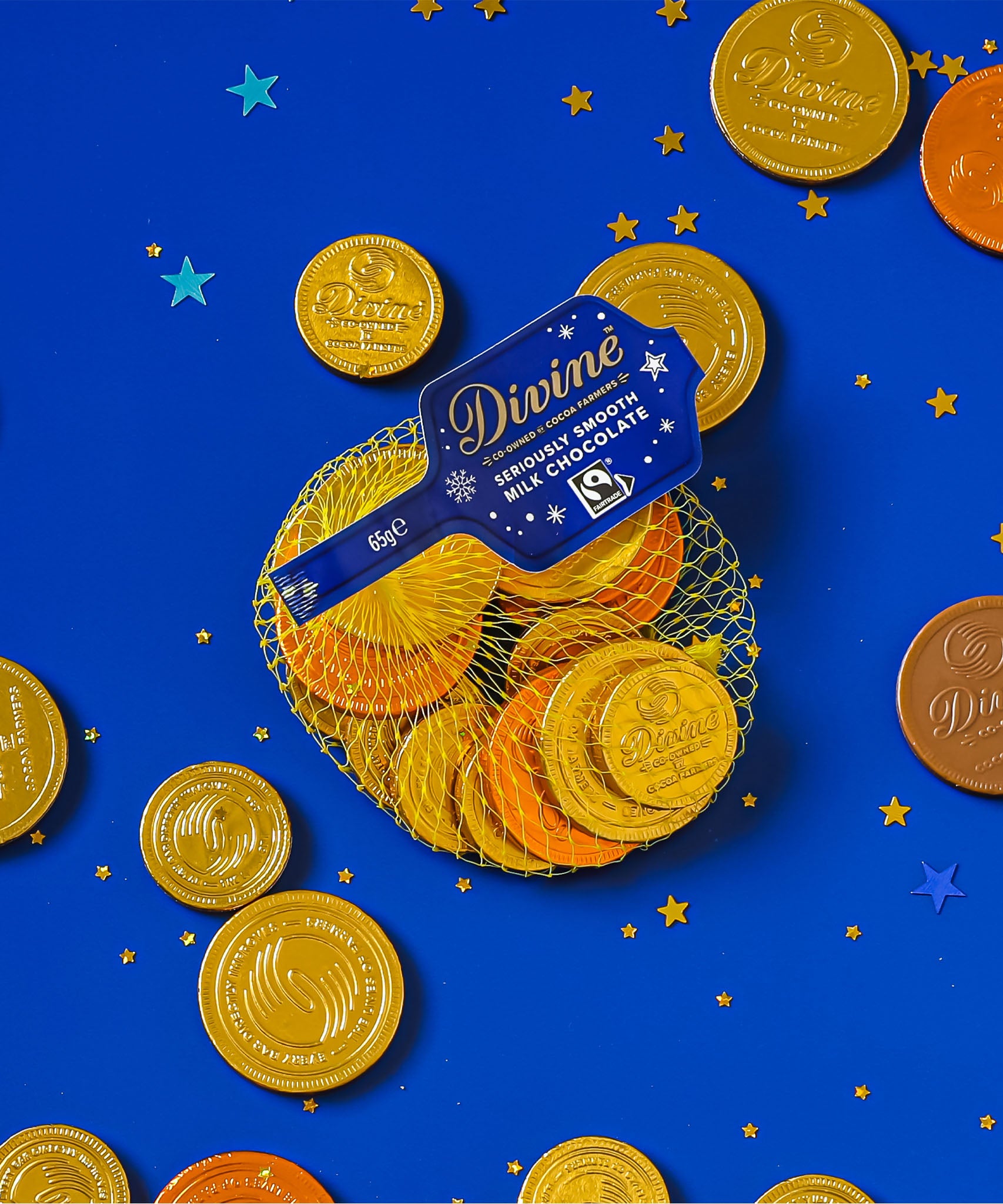 A flatlay with the Divine fairtrade milk chocolate coins inside the yellow netted bag and other coins scattered with a blue festive inspired background. Chocolate coins available at Babipur.