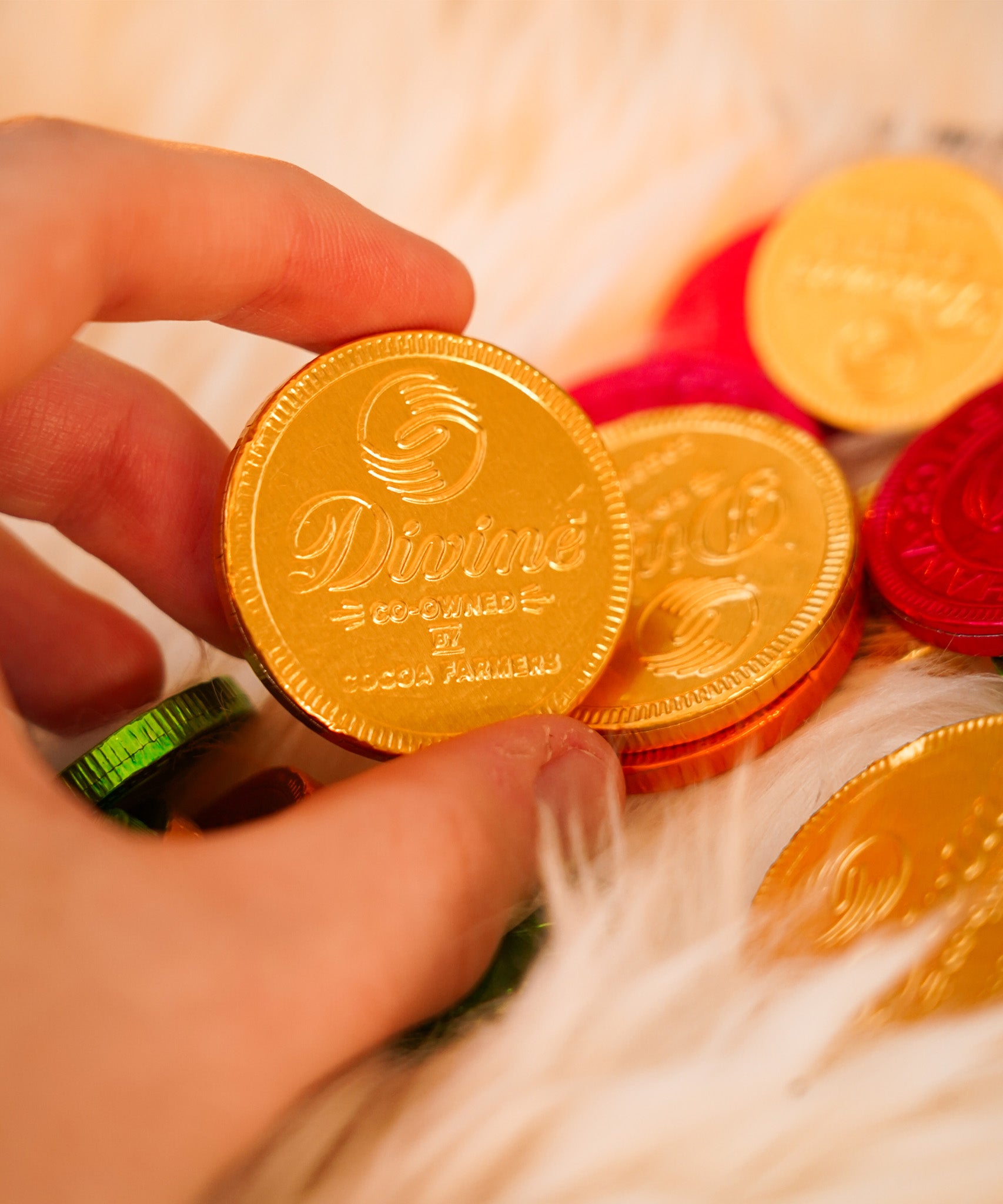 Close up of a hand holding a Divine ethical milk chocolate coin over a fluffy blanket. Coins available at Babipur