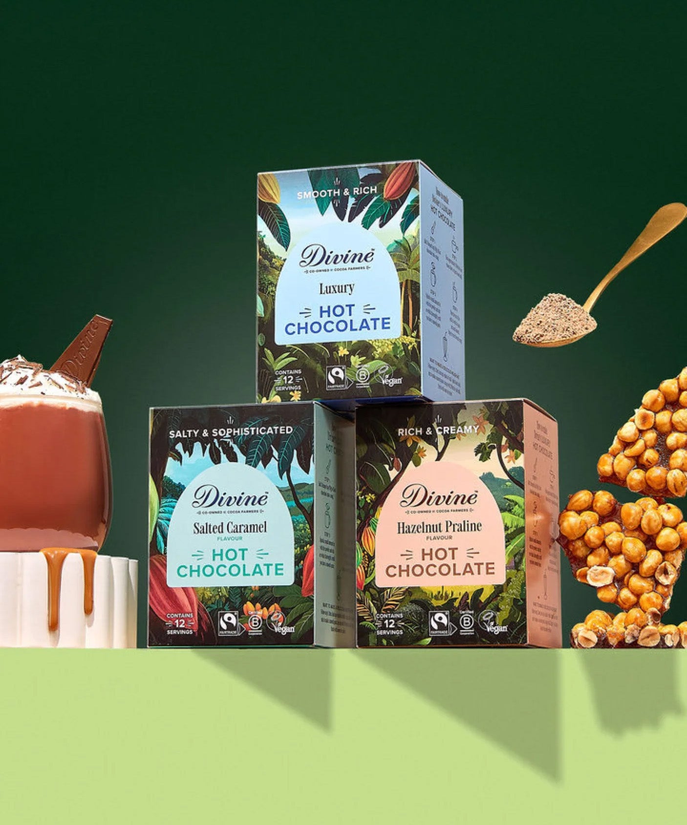 All three Divine fairtrade hot chocolate  boxes displayed on a green platform, shows a golden spoon and a mug of hot chocolate