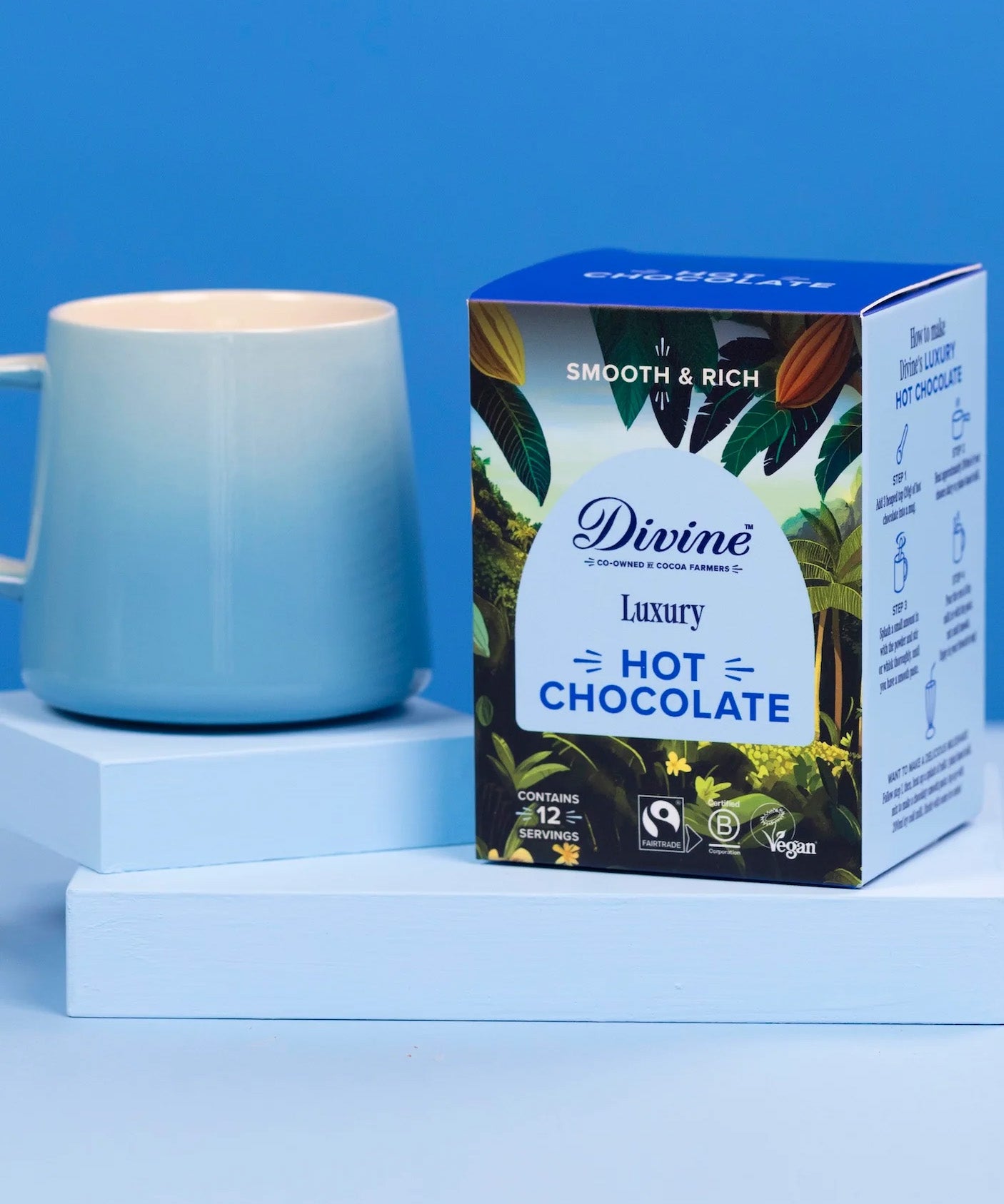 Divine fairtrade luxury hot chocolate box standing on a light blue platform next to a mug.