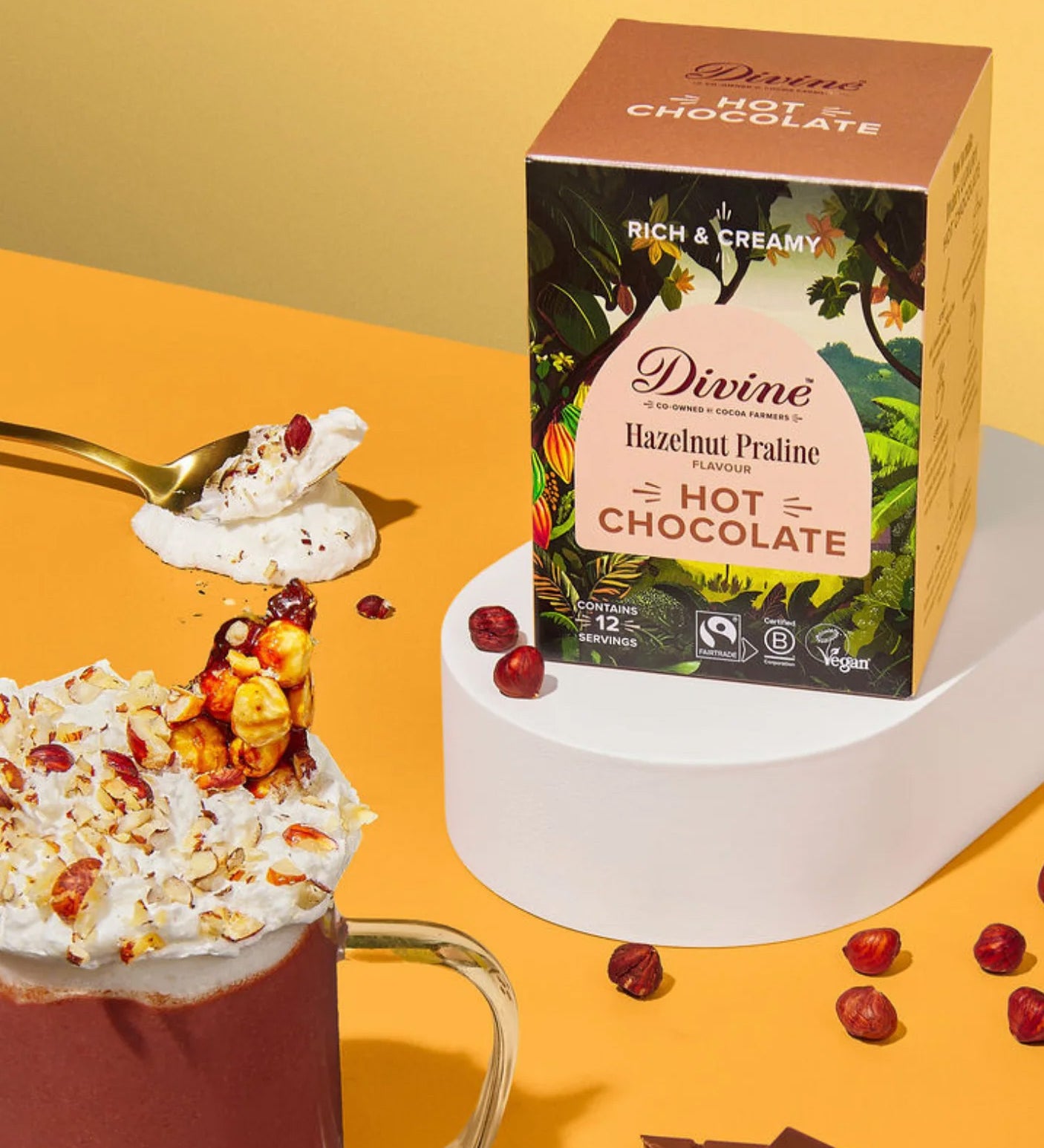 Divine fairtrade luxury hot chocolate with Hazelnut box standing on a white platform next to a mug loose hazelnut pieces