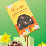 Divine Fairtrade milk chocolate mini eggs with smooth caramel displayed in a decorated yellow and orange box balancing on a green block in between daffodils
