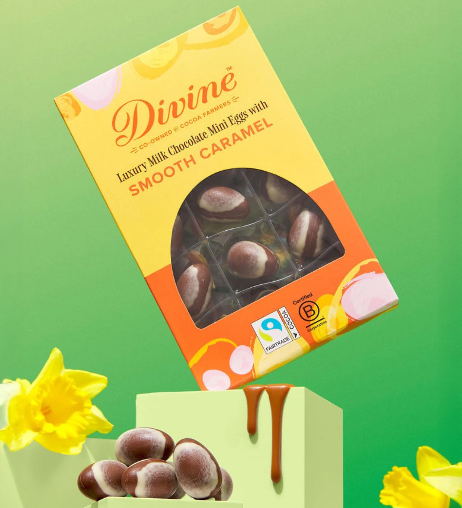 Divine Fairtrade milk chocolate mini eggs with smooth caramel displayed in a decorated yellow and orange box balancing on a green block in between daffodils
