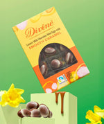 Divine Fairtrade milk chocolate mini eggs with smooth caramel displayed in a decorated yellow and orange box balancing on a green block in between daffodils