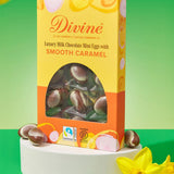 Divine Fairtrade milk chocolate mini eggs with smooth caramel displayed in a decorated yellow and orange box standing on a white block next to daffodils
