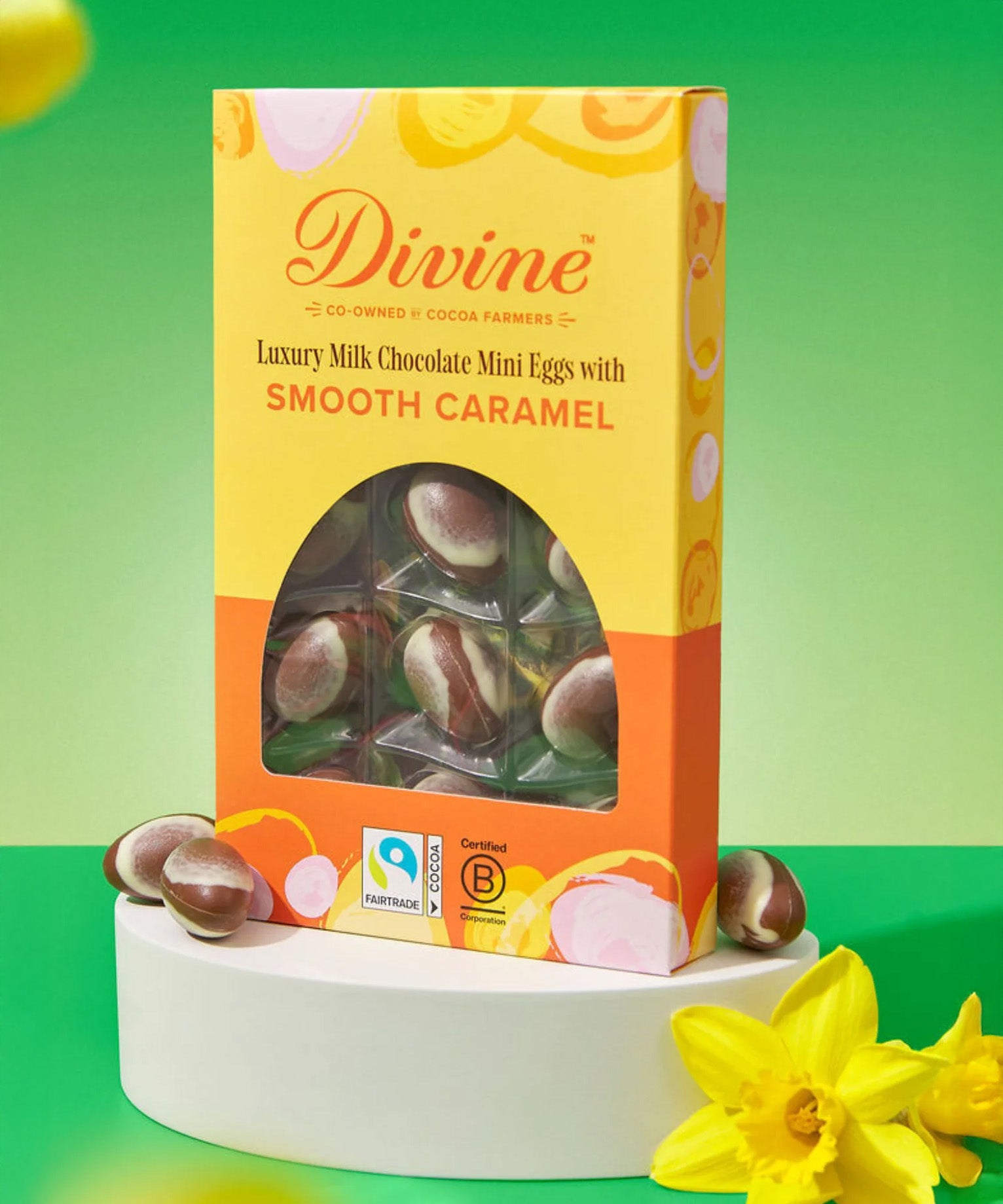 Divine Fairtrade milk chocolate mini eggs with smooth caramel displayed in a decorated yellow and orange box standing on a white block next to daffodils