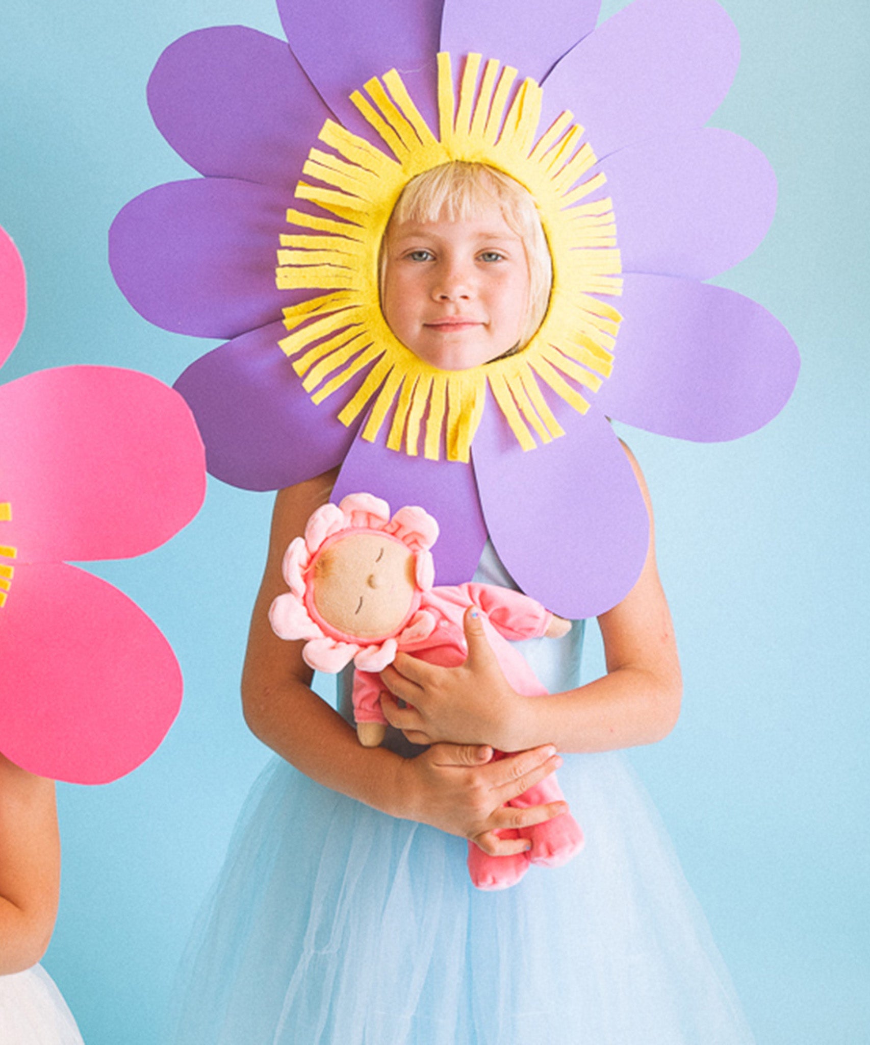 Child holding Olli Ella Dozy Dinkum Doll in Fuchsia Twinkle, wearing a soft pink velour onesie with a flower-shaped hood, available at Babipur.