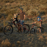A parents with children taking a bike ride through a grassy area. A child behind the parent is riding the Early Rider Seeker X16 16" Bike - Brushed Aluminium