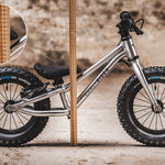 The Early Rider Big Foot Balance Bike stood in front of a light brown wooden ruler, showing the height from the floor to the top of the black seat