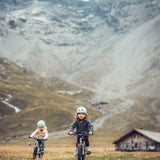 Two children riding bicycles towards the camera, in a mountainous landscape with a wooden cabin In the background.
