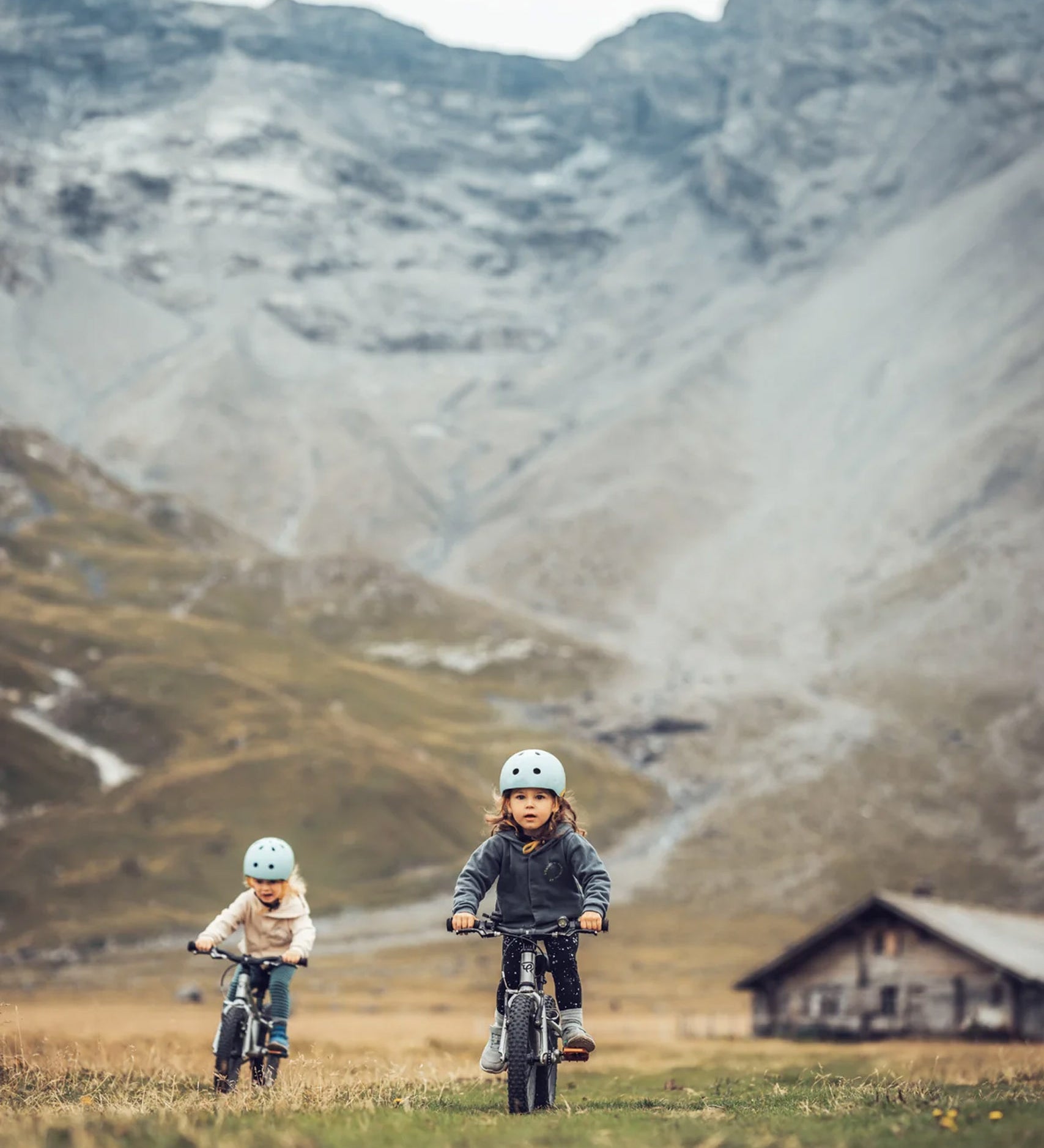 Two children riding bicycles towards the camera, in a mountainous landscape with a wooden cabin In the background.