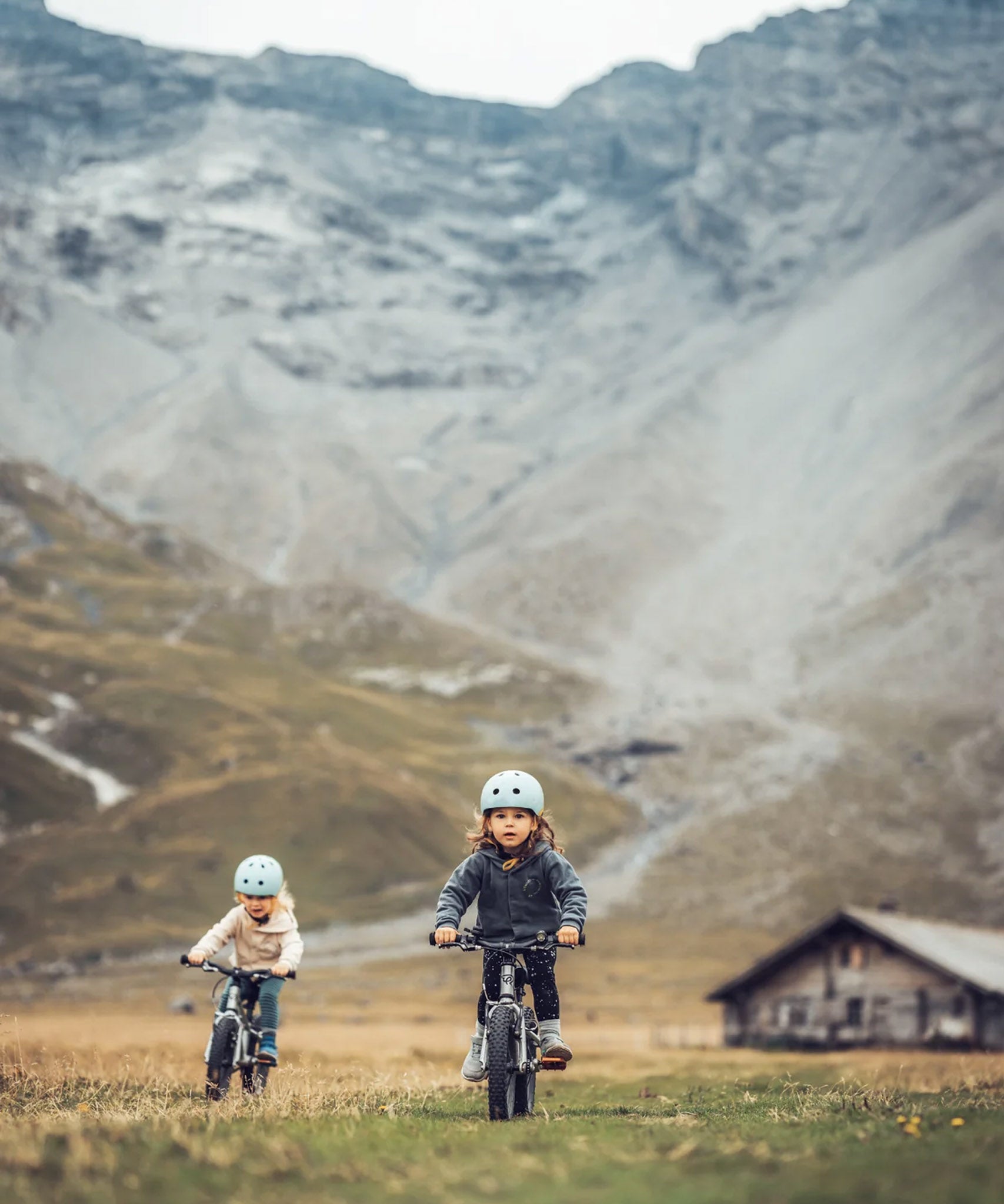 Two children riding bicycles towards the camera, in a mountainous landscape with a wooden cabin In the background.