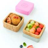 Two open bamboo snack pots, one holding biscuits, the other melon balls with the pink lid above and white lid beneath the pot. 