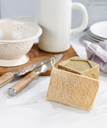 A close up of the Ecoliving loofah kitchen sponge scrubber available at Babipur showing the loofah side of the sponge proped up against a Ecoliving dish soap with other utensils in the background
