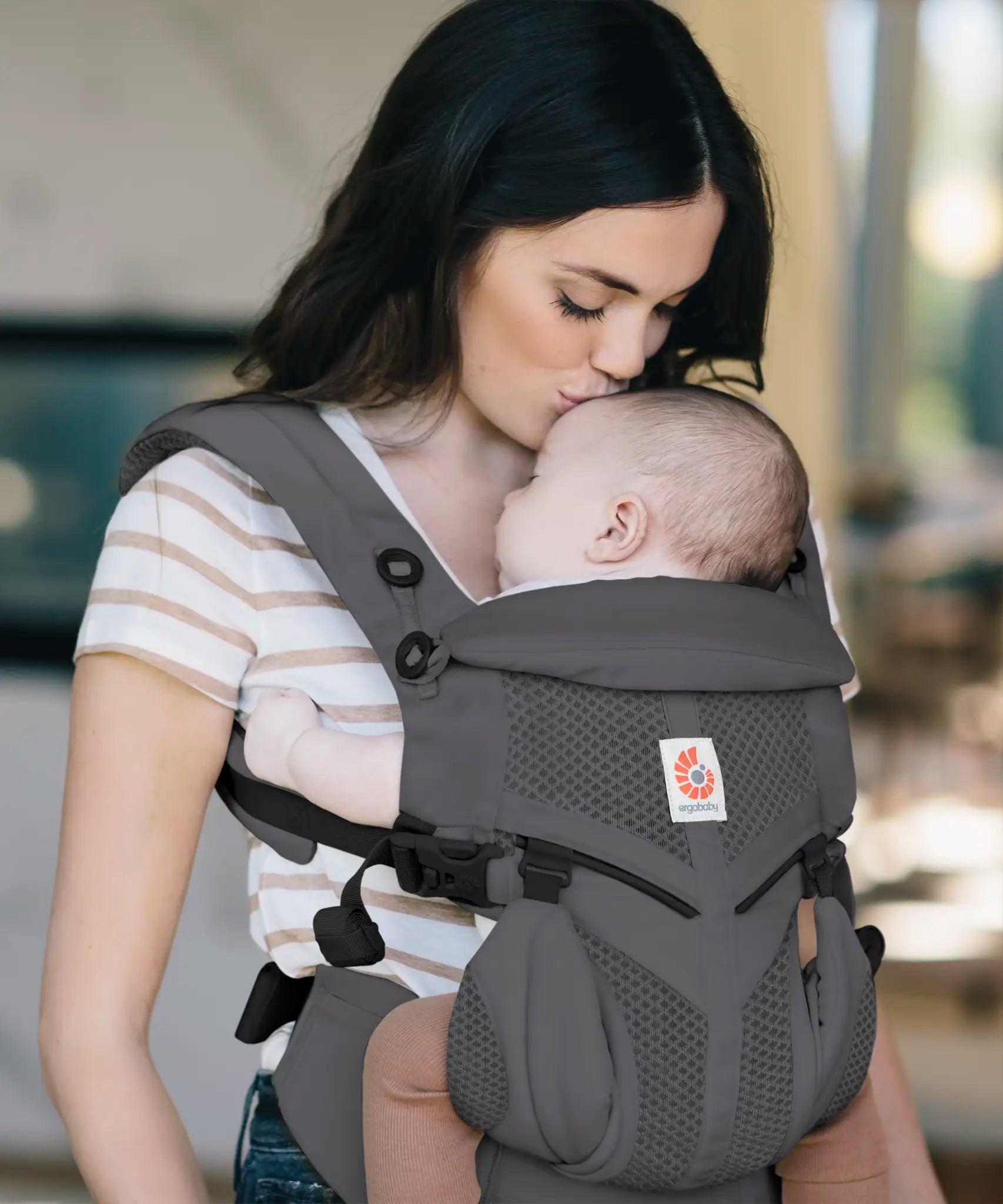 Mother carrying baby in a charcoal grey Ergobaby omni 360 cool air mesh baby carrier and kissing baby's head