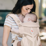 Mother carrying baby in a natural beige Ergobaby omni 360 cool air mesh baby carrier and kissing baby's head