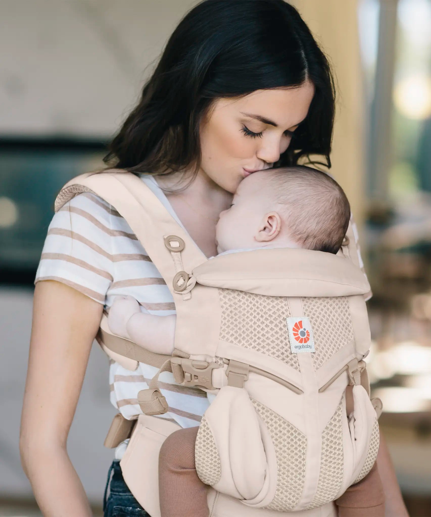 Mother carrying baby in a natural beige Ergobaby omni 360 cool air mesh baby carrier and kissing baby's head