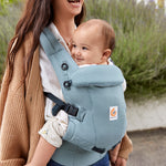 Woman with long brown hair, laughing, standing in a courtyard wearing her baby in a Ergobaby Adapt  SoftTouch™  Cotton Baby Carrier in Slate Blue.
