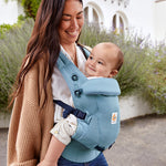 Woman with long brown hair standing in a courtyard wearing her baby in a Ergobaby Adapt  SoftTouch™  Cotton Baby Carrier in Slate Blue.