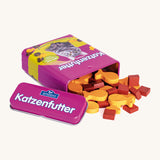 A open purple Erzi cat food tin lying flat next to the lid with the different shaped cat food wooden pieces pouring out of the tin.